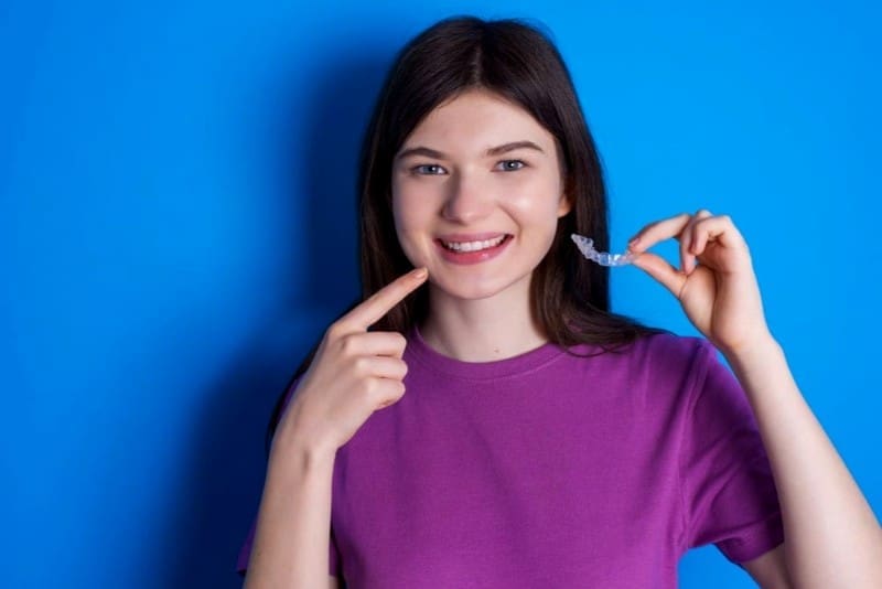 young woman pointing at her teeth smiling and holding an Invisalign clear aligner from Levine Orthodontics, an orthodontist in Cape Coral and Fort Myers, FL